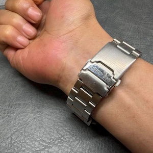 May include: A silver-toned metal watch band featuring a brushed finish and a fold-over clasp mechanism. The clasp is engraved with the text "HI-KRAFT" and includes a secure locking latch, showcasing a standard link design with a metallic sheen.