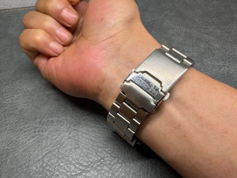 May include: A silver-toned metal watch band featuring a brushed finish and a fold-over clasp mechanism. The clasp is engraved with the text "HI-KRAFT" and includes a secure locking latch, showcasing a standard link design with a metallic sheen.
