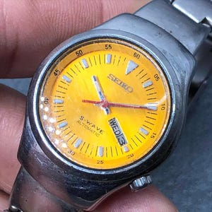 Op de afbeelding: Een vintage Seiko S-Wave automatische polshorloge met een felgele wijzerplaat. Het horloge heeft een zilverkleurige metalen band en kast. De wijzerplaat toont de dag van de week en tijdmarkeringen. De wijzers zijn zilver en rood.