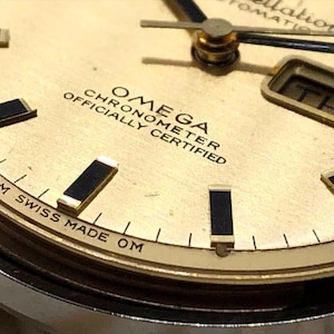 Puede incluir: Primer plano de una esfera de reloj Omega Constellation dorada. El reloj presenta marcadores de hora, agujas y la visualizaci&oacute;n del d&iacute;a de la semana en negro y dorado. El texto "OMEGA CHRONOMETER OFFICIALLY CERTIFIED" es visible. El reloj est&aacute; marcado "OM SWISS MADE OM".