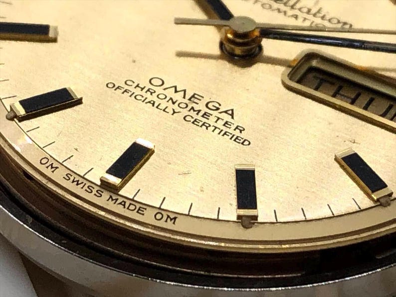 Puede incluir: Primer plano de una esfera de reloj Omega Constellation dorada. El reloj presenta marcadores de hora, agujas y la visualizaci&oacute;n del d&iacute;a de la semana en negro y dorado. El texto "OMEGA CHRONOMETER OFFICIALLY CERTIFIED" es visible. El reloj est&aacute; marcado "OM SWISS MADE OM".