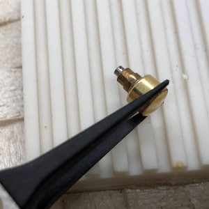 May include: A close-up of a small, brass-colored object held by black tweezers. The object has a cylindrical shape with a threaded end and a flat, circular top. The background is a white, ridged surface.