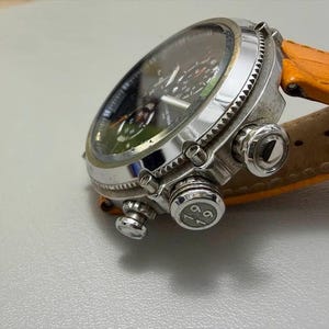 以下が含まれることがあります： 黒い文字盤と複数のダイヤルを備えたシルバーの腕時計。 時計にはテクスチャード加工されたベゼルと明るいオレンジ色の革バンドが付いています。 時計の文字盤には緑と白の詳細が表示されます。 時計は角度が付けられており、側面と上面が表示されています。