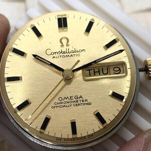 Puede incluir: Esfera de reloj Omega Constellation dorada con agujas e indicadores negros. La esfera muestra el d&iacute;a y la fecha, con el texto "Constellation AUTOMATIC" y "OMEGA CHRONOMETER OFFICIALLY CERTIFIED". El reloj tiene una corona dorada.