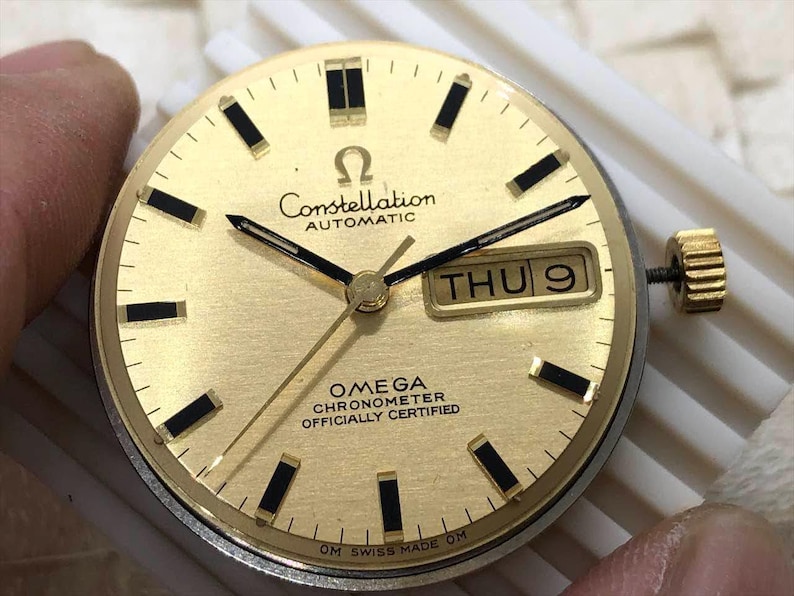 Puede incluir: Esfera de reloj Omega Constellation dorada con agujas e indicadores negros. La esfera muestra el d&iacute;a y la fecha, con el texto "Constellation AUTOMATIC" y "OMEGA CHRONOMETER OFFICIALLY CERTIFIED". El reloj tiene una corona dorada.