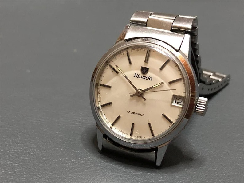 Puede incluir: Reloj Nivada vintage con caja y correa plateadas. La esfera crema tiene marcadores y agujas negras. El reloj tiene una visualizaci&oacute;n de fecha a las 3 en punto y el texto "Nivada" est&aacute; impreso en la esfera.