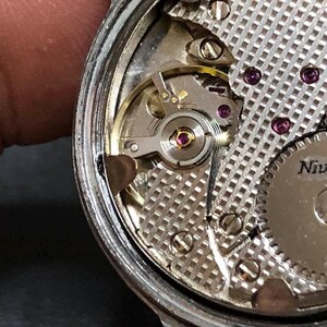 Puede incluir: Primer plano del movimiento de un reloj Nivada. El intrincado mecanismo presenta engranajes plateados, rub&iacute;es y una placa plateada texturizada. Los componentes internos del reloj son visibles, mostrando la precisi&oacute;n del reloj.