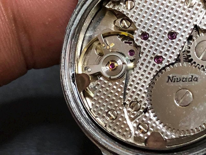Puede incluir: Primer plano del movimiento de un reloj Nivada. El intrincado mecanismo presenta engranajes plateados, rub&iacute;es y una placa plateada texturizada. Los componentes internos del reloj son visibles, mostrando la precisi&oacute;n del reloj.