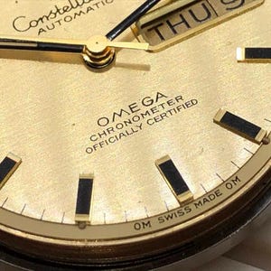 Puede incluir: Primer plano de una esfera de reloj autom&aacute;tico Omega Constellation dorada. El reloj presenta agujas negras y doradas, marcadores de hora rectangulares negros y una pantalla de d&iacute;a y fecha. El texto "OMEGA CHRONOMETER OFFICIALLY CERTIFIED" es visible.