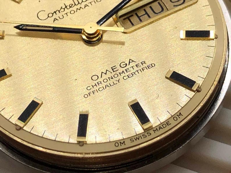 Puede incluir: Primer plano de una esfera de reloj autom&aacute;tico Omega Constellation dorada. El reloj presenta agujas negras y doradas, marcadores de hora rectangulares negros y una pantalla de d&iacute;a y fecha. El texto "OMEGA CHRONOMETER OFFICIALLY CERTIFIED" es visible.