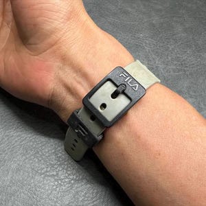 May include: A close-up of a wristwatch with a gray band and a black buckle. The buckle has the brand name "FILA" printed on it. The watch is fastened on a person's wrist.