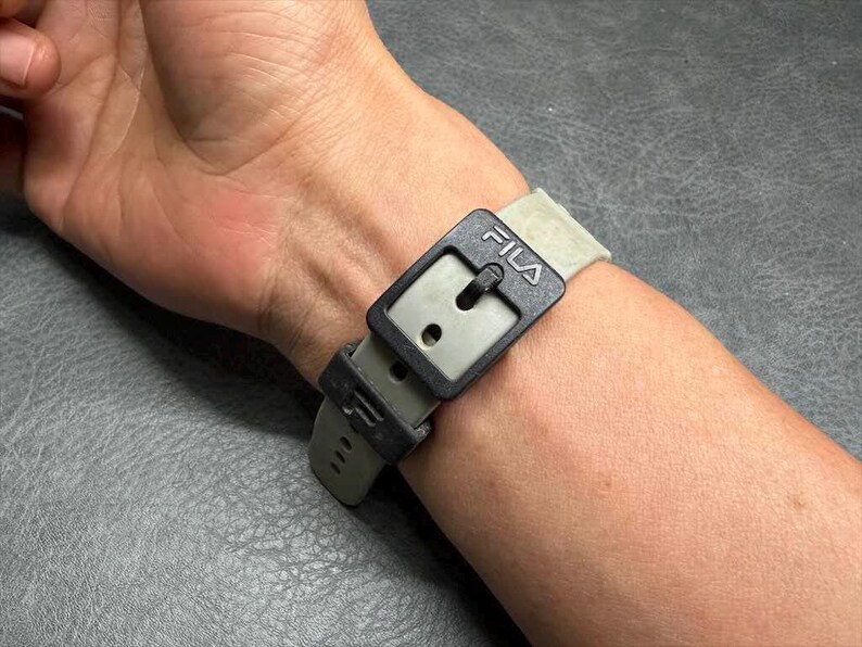 May include: A close-up of a wristwatch with a gray band and a black buckle. The buckle has the brand name "FILA" printed on it. The watch is fastened on a person's wrist.