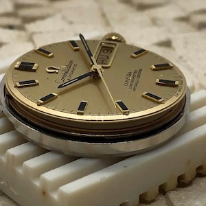 Puede incluir: Una esfera de reloj Omega Constellation Automatic dorada con manecillas e indicadores de hora negros. La esfera muestra el d&iacute;a y la fecha. El reloj est&aacute; sobre una superficie blanca y estriada.