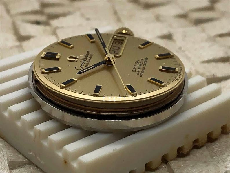 Puede incluir: Una esfera de reloj Omega Constellation Automatic dorada con manecillas e indicadores de hora negros. La esfera muestra el d&iacute;a y la fecha. El reloj est&aacute; sobre una superficie blanca y estriada.