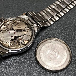Puede incluir: Un reloj Nivada plateado vintage con la parte trasera abierta, que revela los intrincados mecanismos internos. El reloj tiene una correa de metal plateado y una tapa trasera separada. El reloj se muestra sobre una superficie gris oscuro.