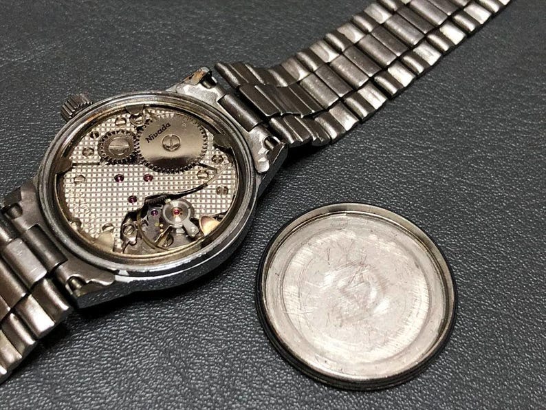 Puede incluir: Un reloj Nivada plateado vintage con la parte trasera abierta, que revela los intrincados mecanismos internos. El reloj tiene una correa de metal plateado y una tapa trasera separada. El reloj se muestra sobre una superficie gris oscuro.