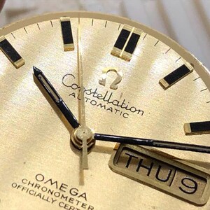 Puede incluir: Primer plano de una esfera de reloj autom&aacute;tico Omega Constellation dorada. El reloj presenta agujas e indicadores de hora negros, una pantalla de d&iacute;a y fecha y el logotipo de Omega. La esfera tiene un acabado texturizado.