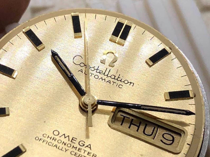 Puede incluir: Primer plano de una esfera de reloj autom&aacute;tico Omega Constellation dorada. El reloj presenta agujas e indicadores de hora negros, una pantalla de d&iacute;a y fecha y el logotipo de Omega. La esfera tiene un acabado texturizado.
