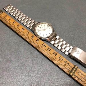 Puede incluir: Un reloj de pulsera vintage con una correa de metal plateado y una esfera blanca con marcadores negros. Una regla de madera con medidas en cent&iacute;metros est&aacute; al lado del reloj. La correa est&aacute; abierta.