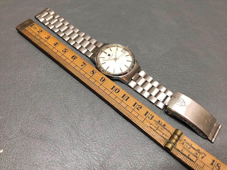Puede incluir: Un reloj de pulsera vintage con una correa de metal plateado y una esfera blanca con marcadores negros. Una regla de madera con medidas en cent&iacute;metros est&aacute; al lado del reloj. La correa est&aacute; abierta.