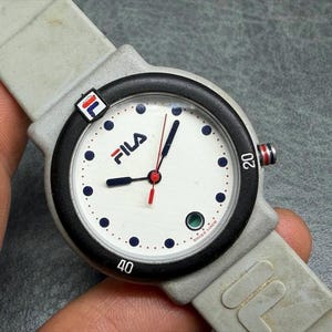 May include: A Fila wristwatch with a white face, black bezel, and gray band. The watch face features the Fila logo, blue dots, and red and blue hands. The watch has a gray band and a small Fila logo on the side.