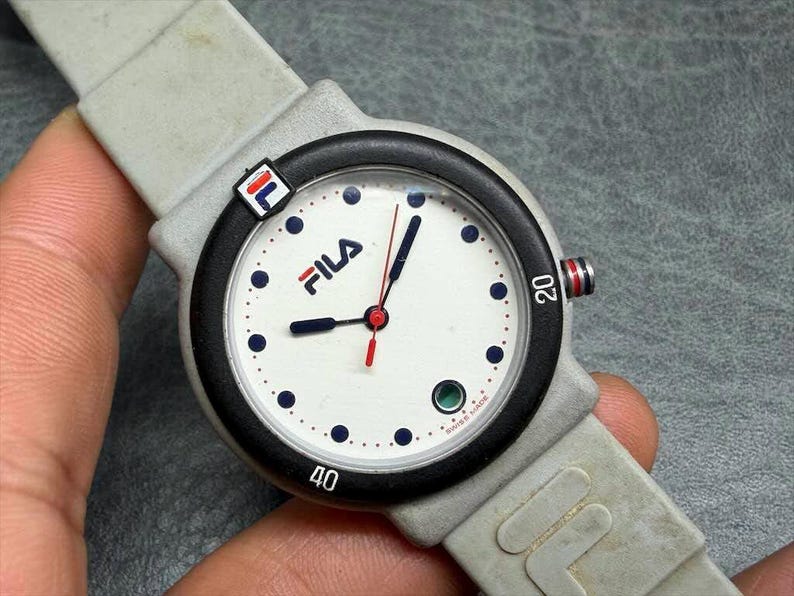 May include: A Fila wristwatch with a white face, black bezel, and gray band. The watch face features the Fila logo, blue dots, and red and blue hands. The watch has a gray band and a small Fila logo on the side.