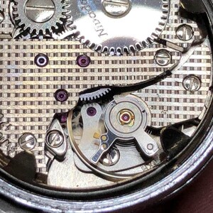 Puede incluir: Primer plano del mecanismo de un reloj, que revela engranajes, tornillos y joyas intrincados. Los componentes plateados est&aacute;n dispuestos sobre un fondo estampado. La precisi&oacute;n del dise&ntilde;o del reloj es evidente.