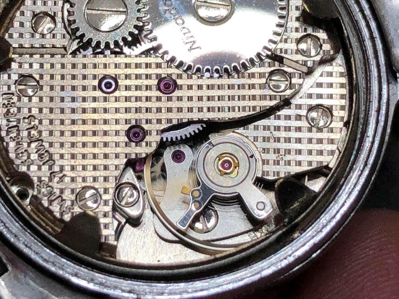 Puede incluir: Primer plano del mecanismo de un reloj, que revela engranajes, tornillos y joyas intrincados. Los componentes plateados est&aacute;n dispuestos sobre un fondo estampado. La precisi&oacute;n del dise&ntilde;o del reloj es evidente.