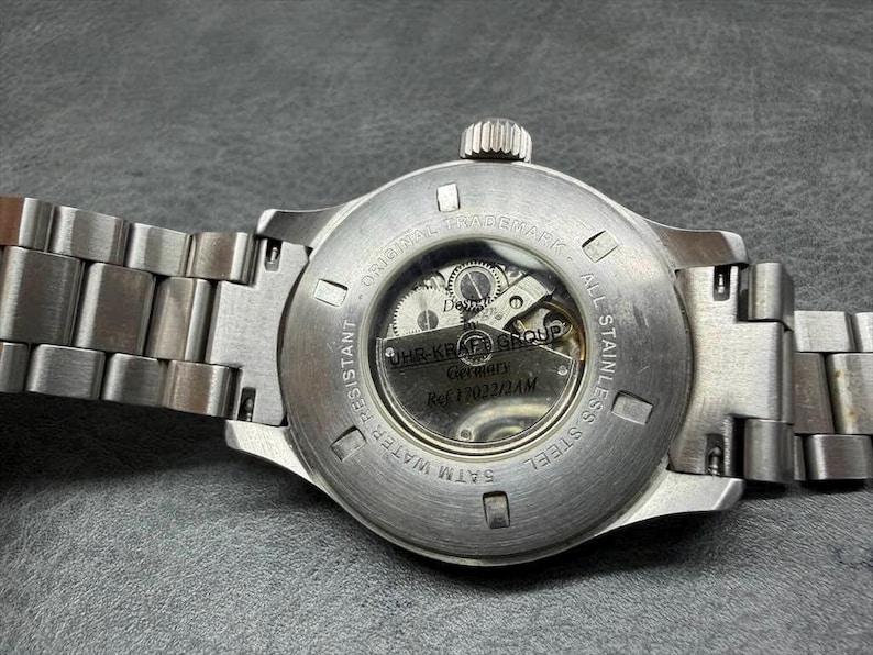 May include: A stainless steel wristwatch with a metal link bracelet, featuring an exhibition case back that reveals the internal mechanical movement. The back is engraved with UHR-KRAFT GROUP, Germany, Ref 17022/2AM, 5ATM WATER RESISTANT.