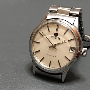 Puede incluir: Reloj Nivada vintage con caja y correa plateadas. La esfera es de color crema con marcadores y agujas negras. El reloj tiene el logotipo de Nivada y el texto "17 JEWELS" y "SWISS MADE". La fecha se muestra a las 3 en punto.