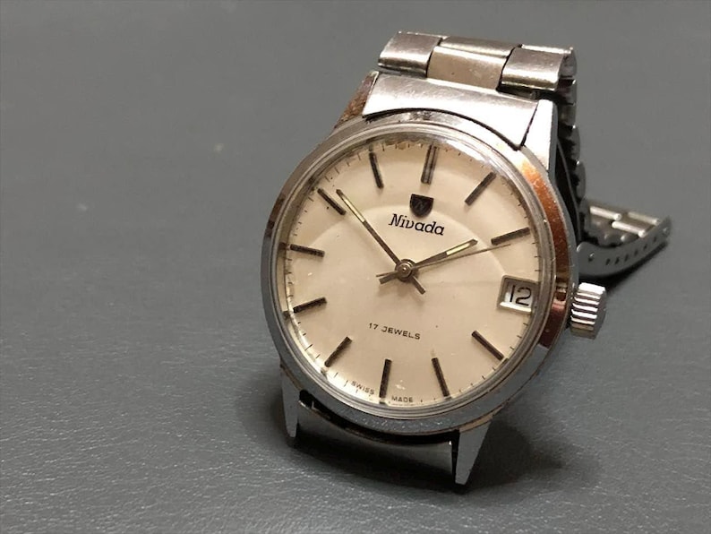 Puede incluir: Reloj Nivada vintage con caja y correa plateadas. La esfera es de color crema con marcadores y agujas negras. El reloj tiene el logotipo de Nivada y el texto "17 JEWELS" y "SWISS MADE". La fecha se muestra a las 3 en punto.