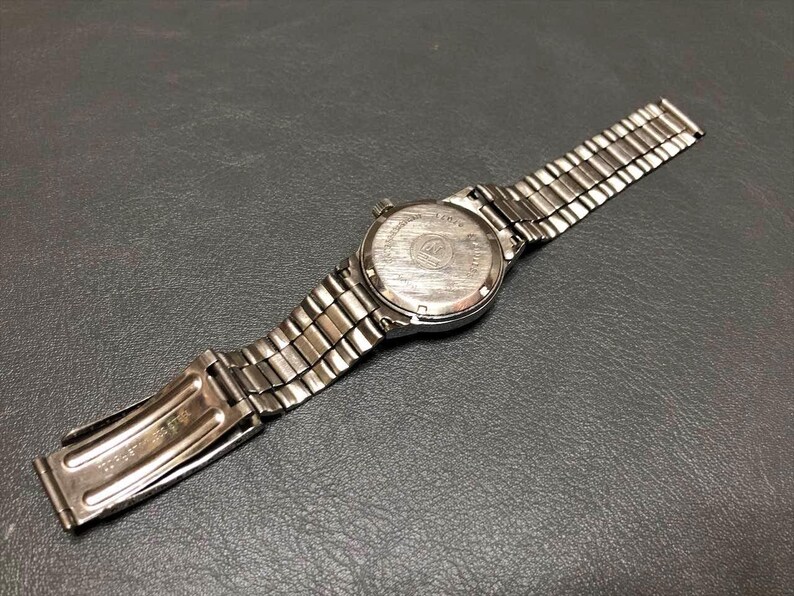Puede incluir: Un reloj de pulsera plateado con una correa de metal, visto desde atr&aacute;s, sobre una superficie oscura. La esfera del reloj es visible y la correa est&aacute; parcialmente abierta. El reloj tiene un dise&ntilde;o cl&aacute;sico y vintage.