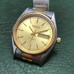 Puede incluir: Un reloj de pulsera automático Tissot vintage con esfera y agujas doradas, y una correa de acero inoxidable plateado. La esfera muestra el día y la fecha, con el día que indica "SUN 16". El reloj tiene un bisel y una corona dorados.