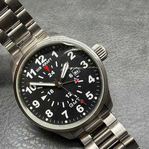 May include: A stainless steel Uir-Kraft wristwatch with a black dial, white Arabic numerals, and a GMT function. The watch features a metal link bracelet, a date window at the three o'clock position, and a red-tipped GMT hand on a black face.