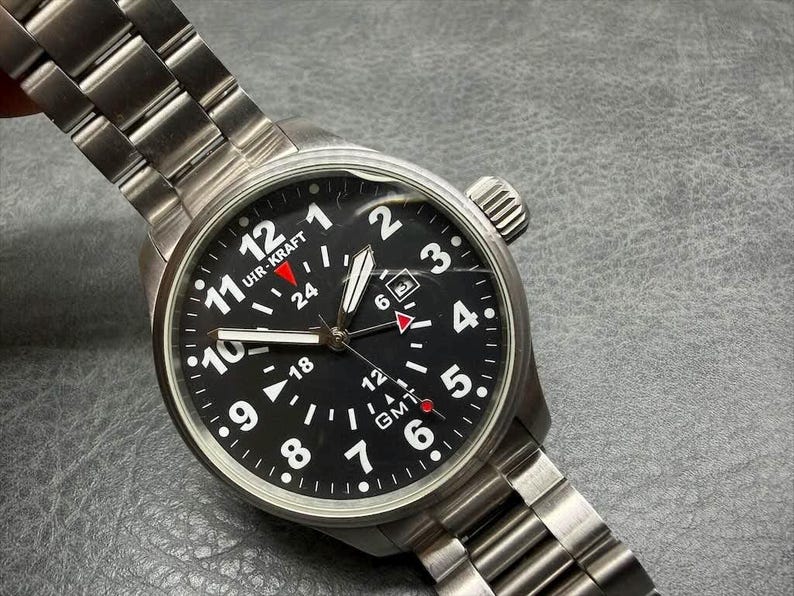 May include: A stainless steel Uir-Kraft wristwatch with a black dial, white Arabic numerals, and a GMT function. The watch features a metal link bracelet, a date window at the three o'clock position, and a red-tipped GMT hand on a black face.