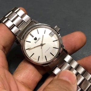 Puede incluir: Un reloj de pulsera Nivada vintage con una correa de metal plateado y una esfera blanca. El reloj presenta marcadores de hora negros, una ventana de fecha a las 3 en punto y la marca "Nivada". El reloj se sostiene en una mano.