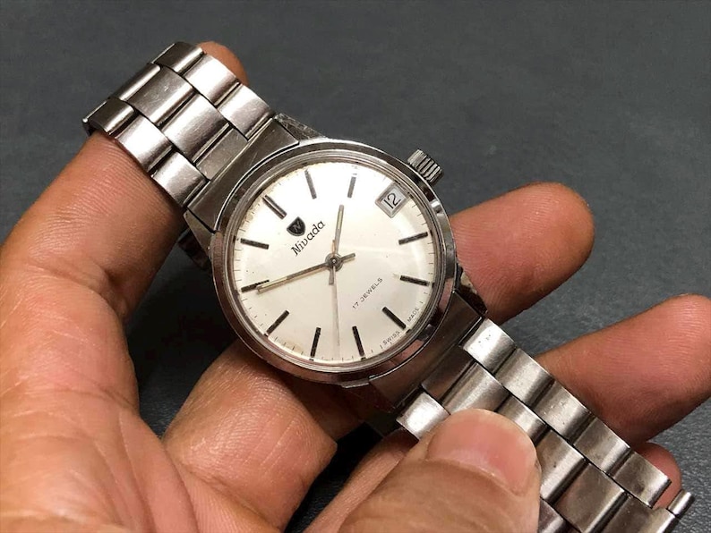 Puede incluir: Un reloj de pulsera Nivada vintage con una correa de metal plateado y una esfera blanca. El reloj presenta marcadores de hora negros, una ventana de fecha a las 3 en punto y la marca "Nivada". El reloj se sostiene en una mano.
