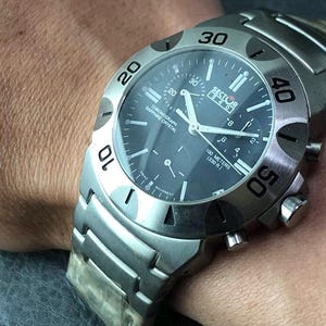 May include: A silver-toned Sector watch with a dark blue face. The watch features a chronograph, sapphire crystal, and is water-resistant to 100 meters (330 ft). The watch has a stainless steel band and a bezel with numerals.