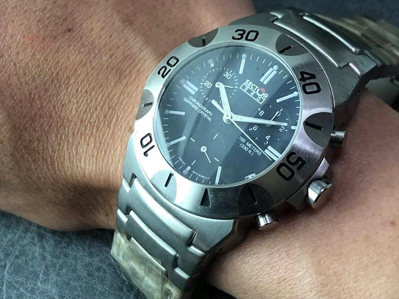 May include: A silver-toned Sector watch with a dark blue face. The watch features a chronograph, sapphire crystal, and is water-resistant to 100 meters (330 ft). The watch has a stainless steel band and a bezel with numerals.