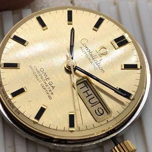 Puede incluir: Una esfera de reloj autom&aacute;tico Omega Constellation dorada con marcadores de hora y agujas rectangulares negros. La pantalla de la fecha muestra "JUE 9". La esfera del reloj tiene el texto "OMEGA CHRONOMETER OFFICIALLY CERTIFIED" y "Constellation AUTOMATIC".