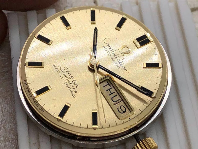Puede incluir: Una esfera de reloj autom&aacute;tico Omega Constellation dorada con marcadores de hora y agujas rectangulares negros. La pantalla de la fecha muestra "JUE 9". La esfera del reloj tiene el texto "OMEGA CHRONOMETER OFFICIALLY CERTIFIED" y "Constellation AUTOMATIC".