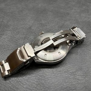 May include: A stainless steel wristwatch shown from the back, featuring a circular case with an exhibition window revealing internal mechanical components. The watch includes a metal link bracelet with a folding deployment clasp mechanism.