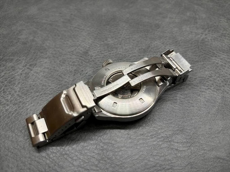May include: A stainless steel wristwatch shown from the back, featuring a circular case with an exhibition window revealing internal mechanical components. The watch includes a metal link bracelet with a folding deployment clasp mechanism.