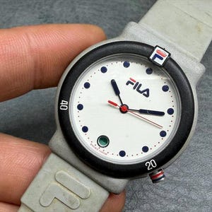 May include: A Fila watch with a white face, black bezel, and gray strap. The watch face features the Fila logo, blue dots, and black hands with a red second hand. The watch has a gray strap with the Fila logo.