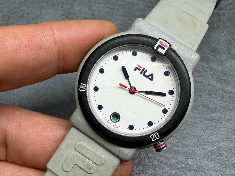 May include: A Fila watch with a white face, black bezel, and gray strap. The watch face features the Fila logo, blue dots, and black hands with a red second hand. The watch has a gray strap with the Fila logo.