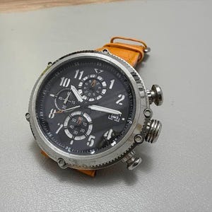 以下が含まれることがあります： 黒い文字盤とオレンジ色の革バンドが付いたステンレス製の腕時計。この時計は、複数のサブダイヤルとシルバーのアクセントが特徴です。この時計は、大きく丸い文字盤に目立つ数字と針が付いています。