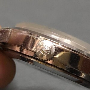 Puede incluir: Primer plano de un reloj de pulsera plateado. El reloj tiene un cristal transparente y abovedado y una corona texturizada. La caja y la correa del reloj parecen ser de metal. El reloj est&aacute; sobre un fondo borroso.