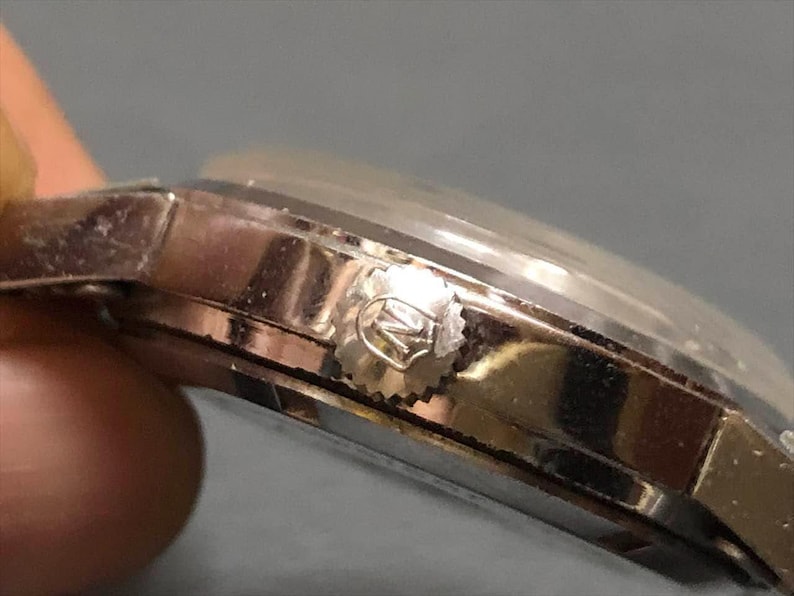 Puede incluir: Primer plano de un reloj de pulsera plateado. El reloj tiene un cristal transparente y abovedado y una corona texturizada. La caja y la correa del reloj parecen ser de metal. El reloj est&aacute; sobre un fondo borroso.
