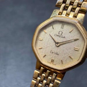May include: A gold-tone Omega De Ville wristwatch featuring an octagonal case and a textured champagne-colored dial. The watch includes gold-tone hour markers, thin hands, and a matching gold-tone metal link bracelet with a polished finish.