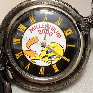 Puede incluir: Un reloj de bolsillo plateado con esfera negra, números romanos amarillos y un personaje de dibujos animados, Piolín. La esfera también incluye el texto "Millennium 2000". El reloj tiene un diseño ornamental alrededor del bisel.